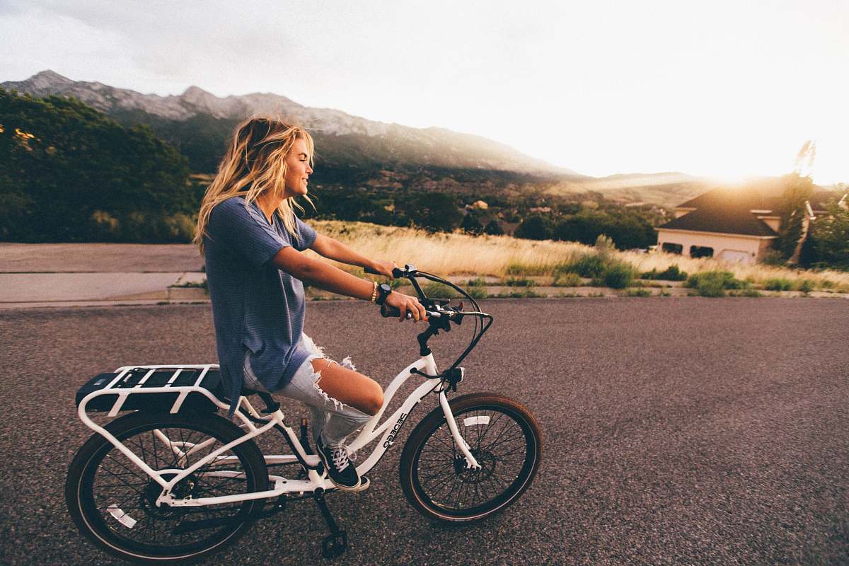 person woman in blue t-shirt and distressed blue denim jeans riding white bicycle on grey road near mountains at daytime human