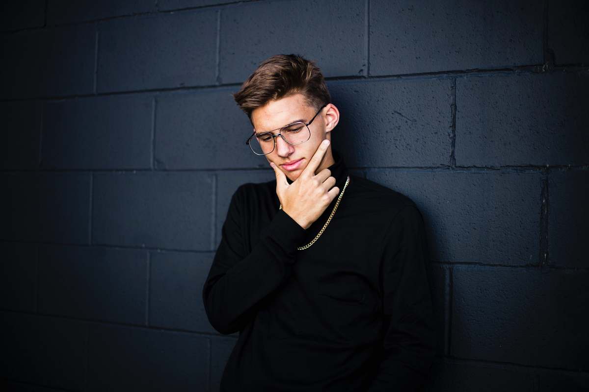 people man leaning on wall while looking down wearing eyeglasses and necklace with right hand on chin human