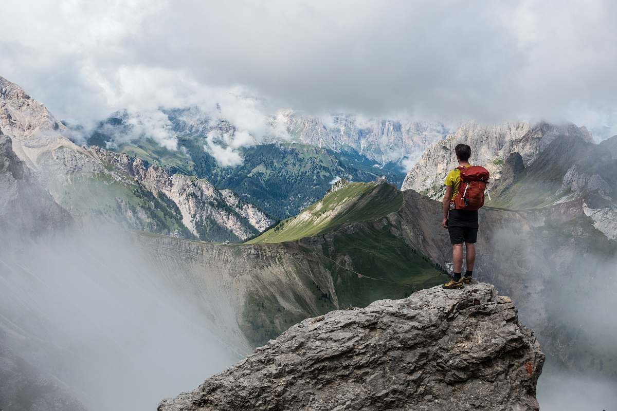 nature man with red backpack standing on cliff facing mountains under white sky during daytime hiking