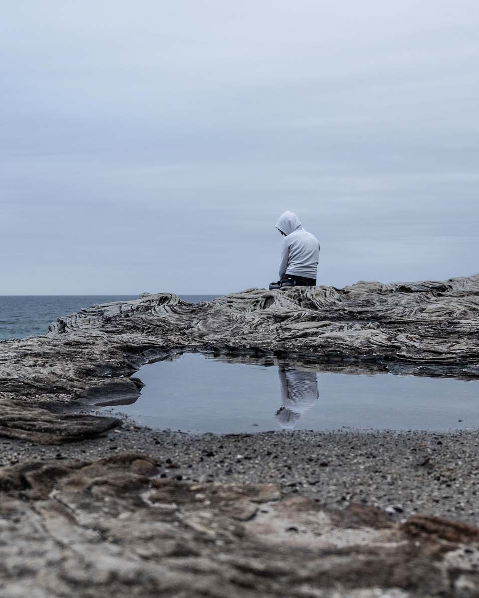 human man sitting on cliff overlooking sea during daytime puddle