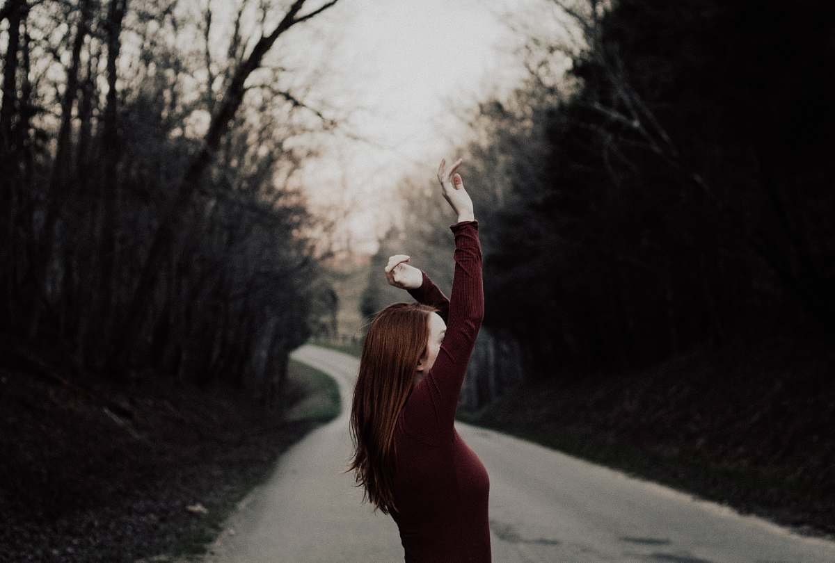 woman selective focus photography of female raising hands in the middle of the road surrounded by withered trees dirt road