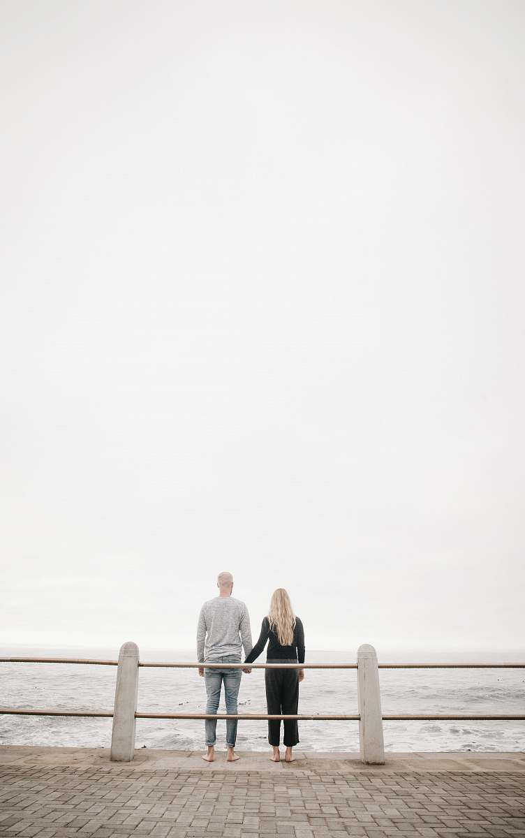 people couple leaning on guard rail while holdings hands facing ocean at daytime person