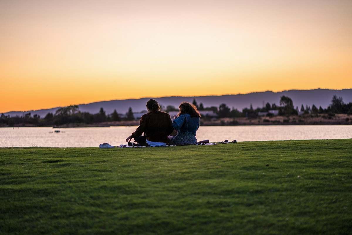 Capture the serene beauty of a couple sitting on lush green grass, gazing lovingly at the tranquil waters as the sun sets in vibrant hues of orange and pink. This picturesque scene evokes feelings of romance and connection, making it perfect for projects related to love, nature, or outdoor activities. Ideal for use in advertisements, blogs, or social media posts celebrating relationships and the beauty of nature.