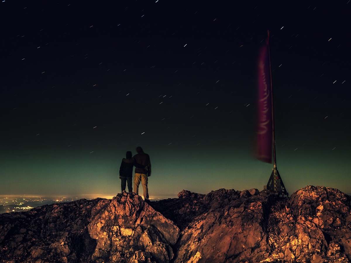 night silhouette of two people standing on brown rocks under starry night stars