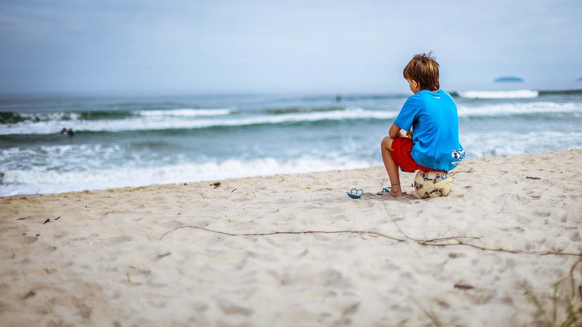 water boy sitting on ball while staring at the ocean beach