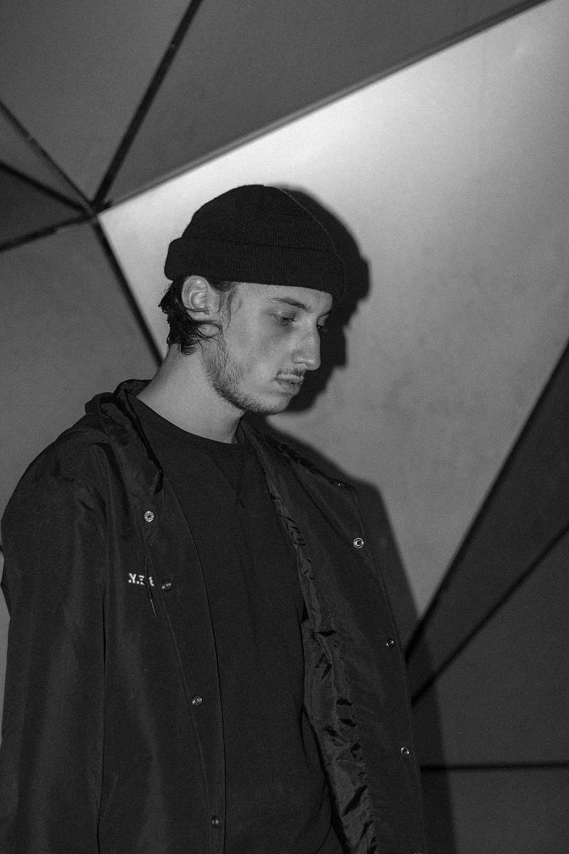 clothing grayscale photography of man wearing jacket and hat standing while facing his right side and looking down apparel