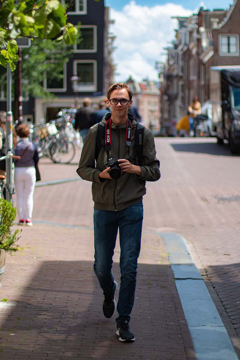 clothing man holding black DSLR camera standing on sidewalk during daytime footwear