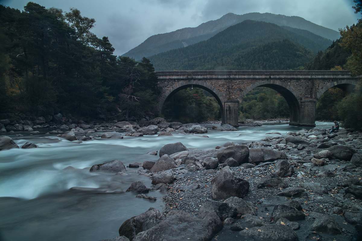 rock gray arch bridge over river under gray sky at daytime mountains