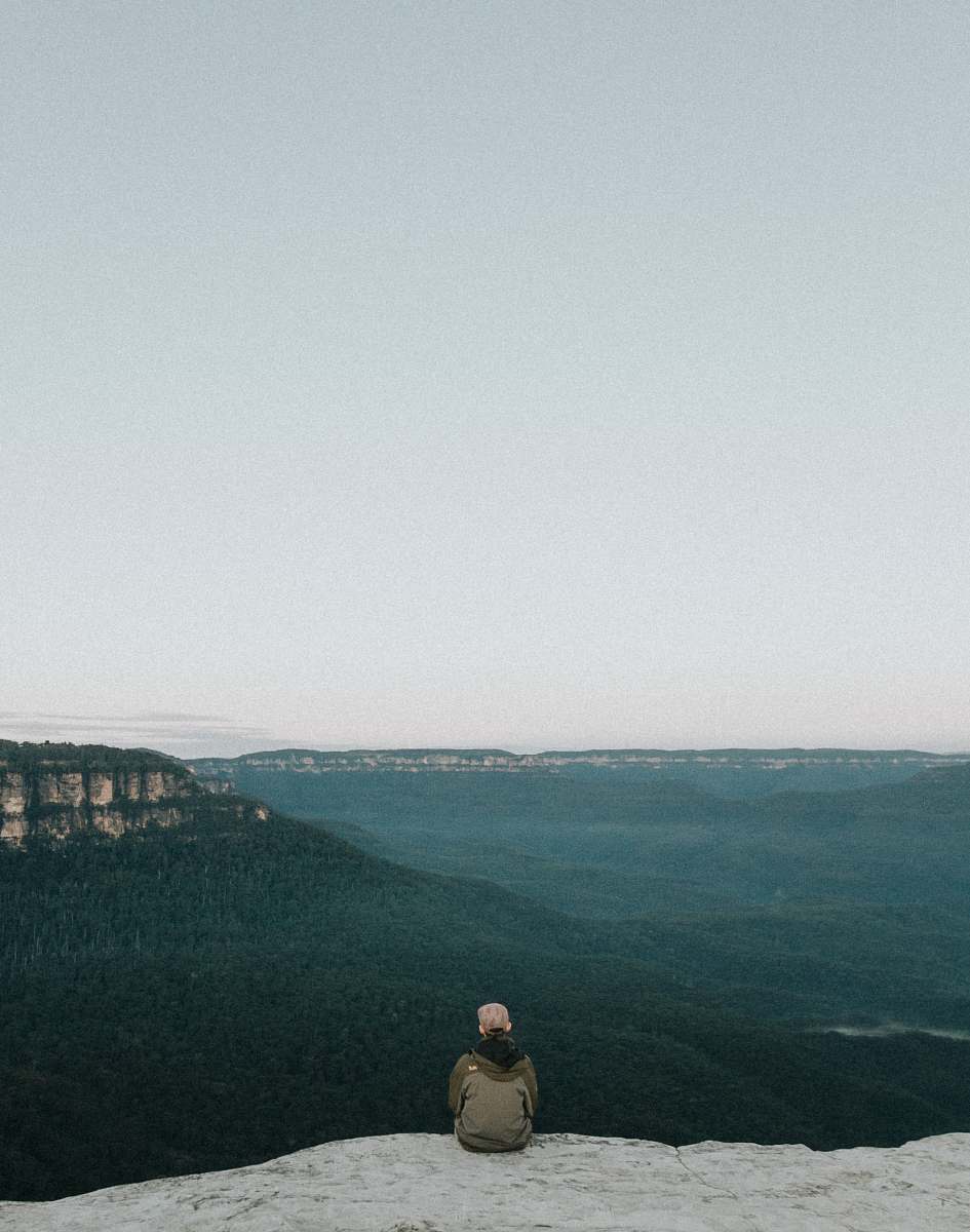 grey person sitting on rock while sight seeing blue mountains