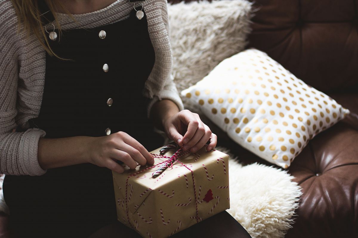 woman in grey sweater and blue denim dungarees sitting on brown and white sofa opening a yellow gift box closeup photography