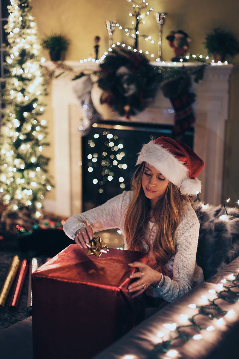 woman putting ribbon in red gift box near lighted Christmas tree inside room