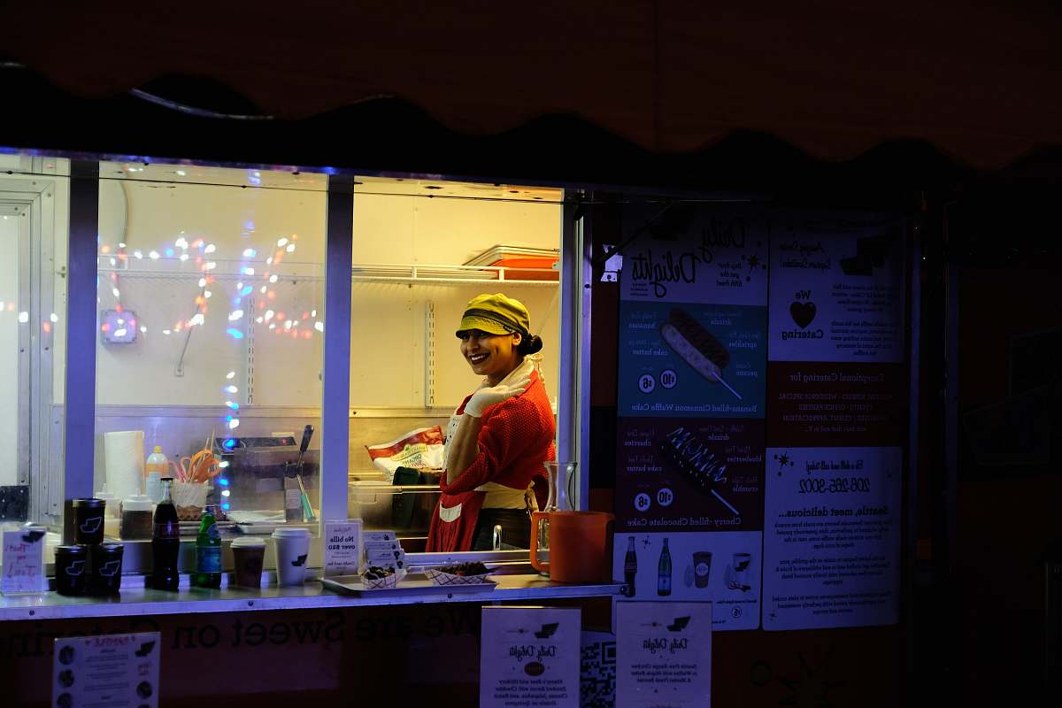 Capture the vibrant atmosphere of an urban night as a man stands at a food stand, with a stunning backdrop of city lights. In the foreground, a woman in a striking red top adds a pop of color to this lively scene. This image is perfect for showcasing nightlife, street food culture, or urban fashion, making it ideal for advertising, blog posts, or social media content.