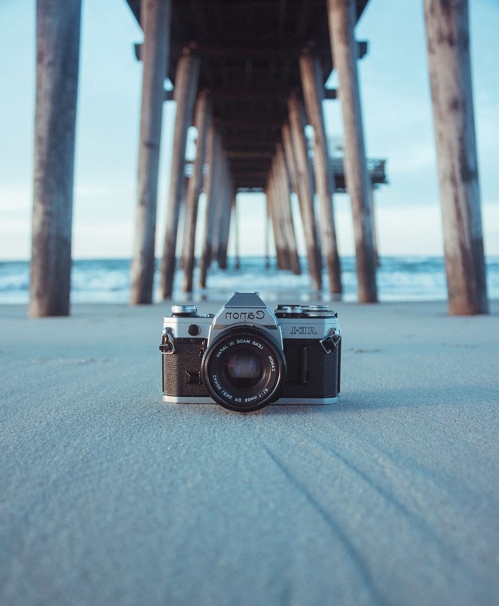 black and gray Canon AE-1 camera on gray sand under brown dock near body of water at daytime