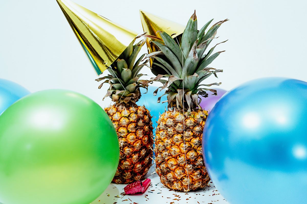 two pineapples with gold party hats near colorful balloons