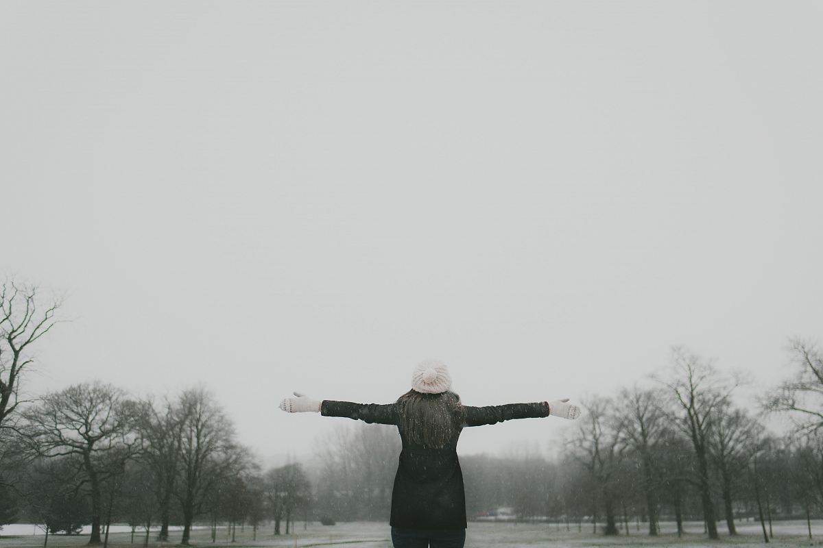 woman in black long-sleeved shirt standing and facing trees with stretched arms under white sky