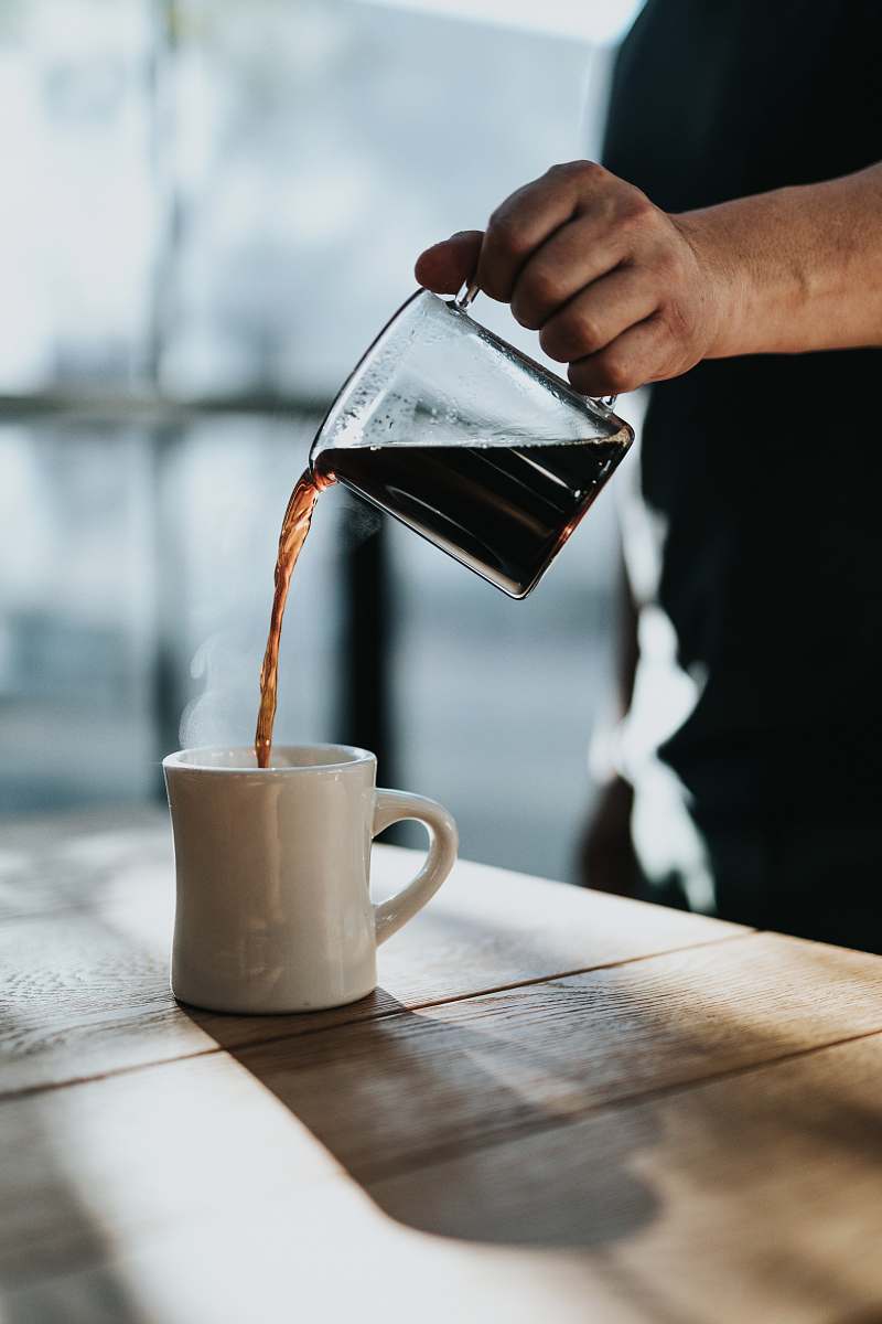 coffee person pouring black coffee in white ceramic mug placed on brown wooden table during daytime person