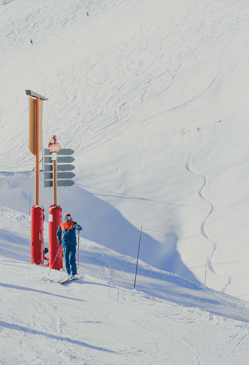 skiing man in blue jacket holding ski poles standing near red and brown post piste