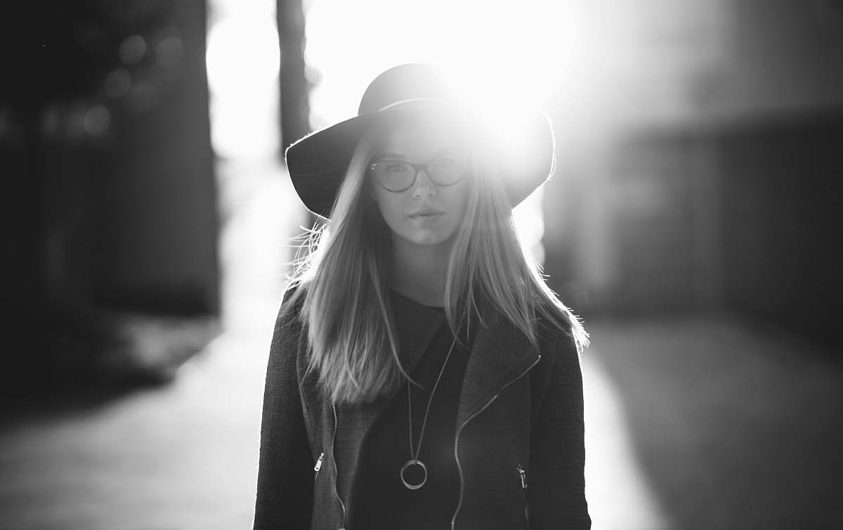 human grayscale photography of woman wearing hat and coat standing near wall female
