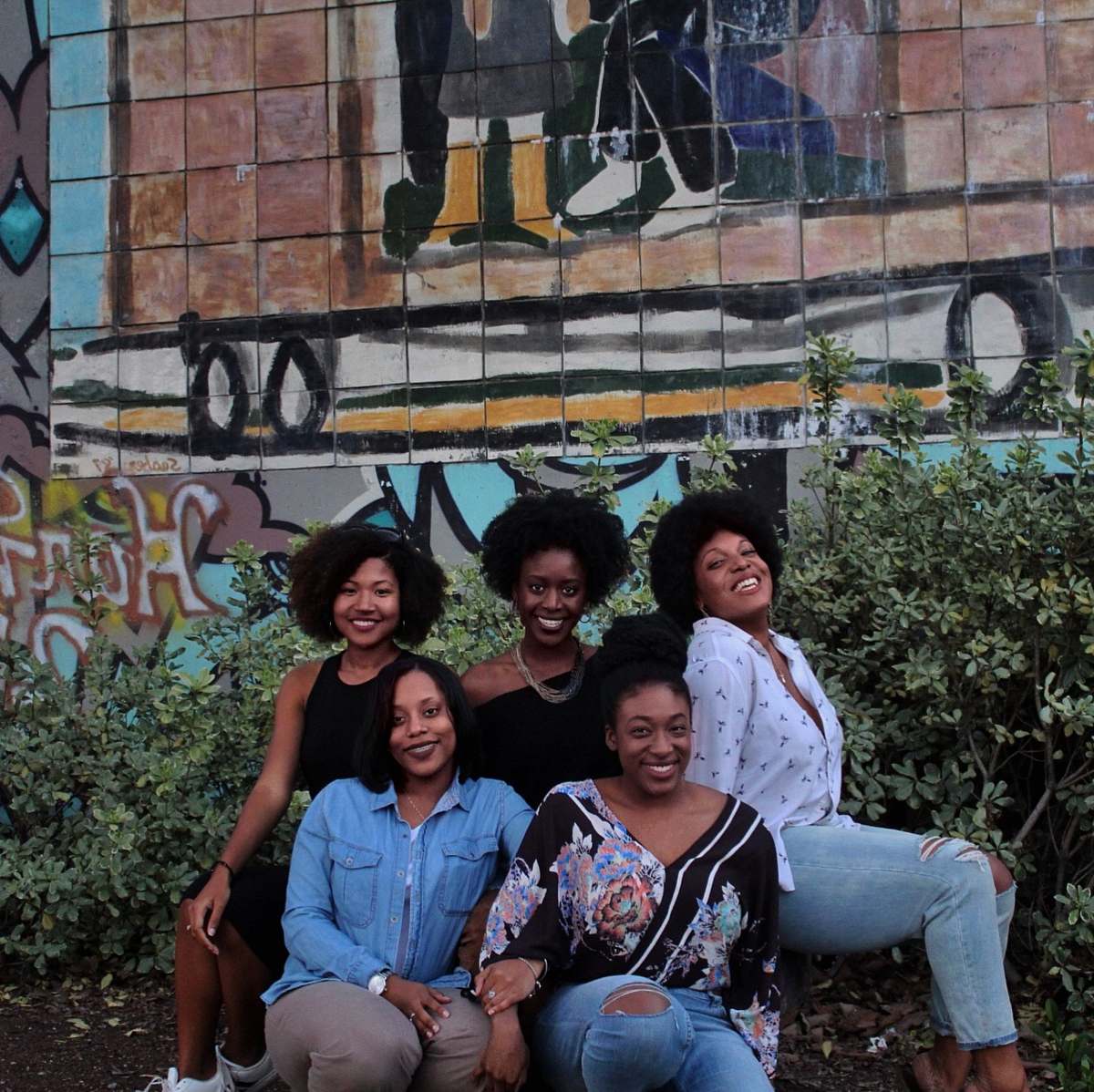This vibrant scene captures five women sharing joyful moments, with three of them seated on a scenic bench in front of a colorful mural. Their smiles radiate happiness and camaraderie, making this image ideal for themes of friendship, community, and outdoor leisure. Perfect for blogs, social media campaigns, or advertisements focused on women's empowerment and social gatherings.