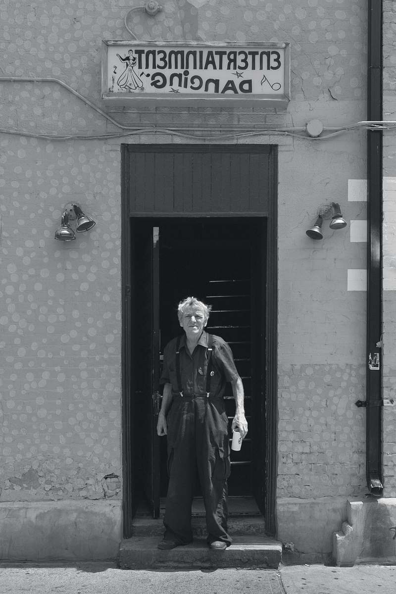 person grayscale photo of man wearing collared tops and dress pants standing between concrete wall near Entertainment Dancing signage building black-and-white