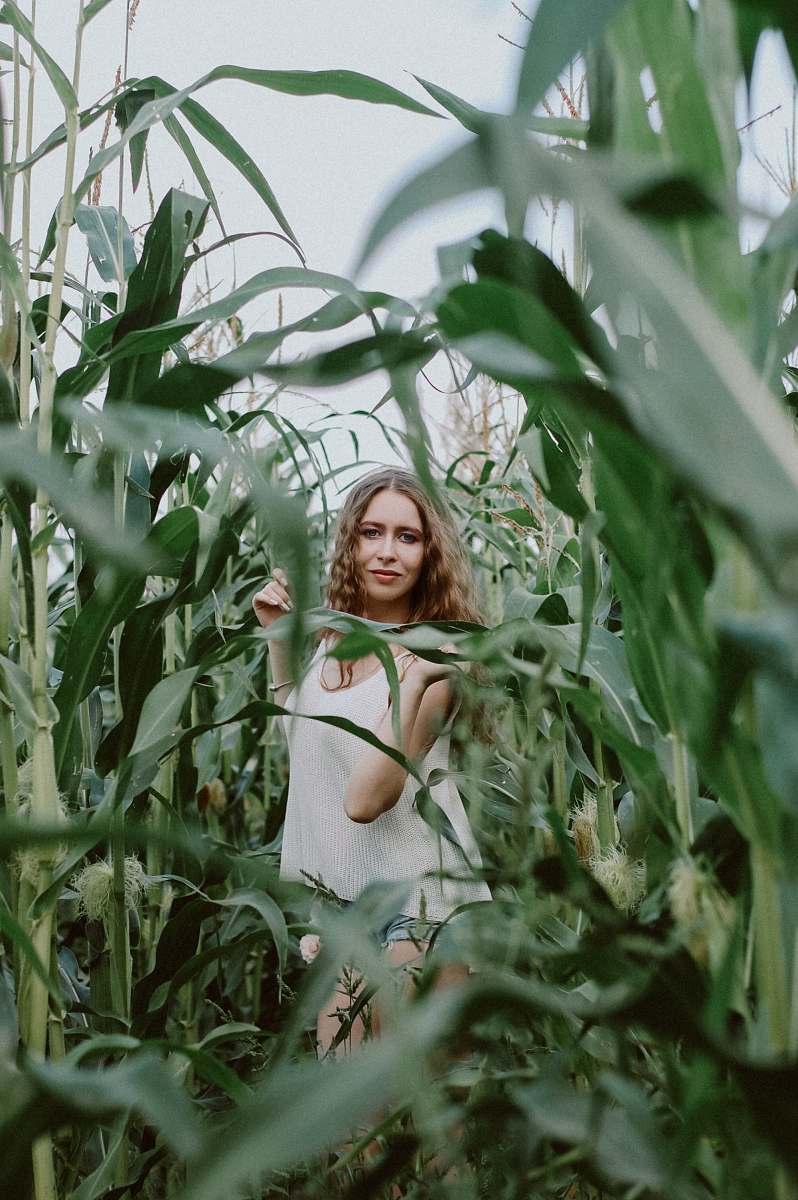 A vibrant and cheerful woman stands confidently in the heart of a sprawling cornfield, surrounded by towering green stalks swaying gently in the breeze. Her genuine smile radiates warmth and positivity, capturing the essence of connection with nature. This image is perfect for agricultural themes, wellness campaigns, and lifestyle blogs focused on rural living or natural beauty.