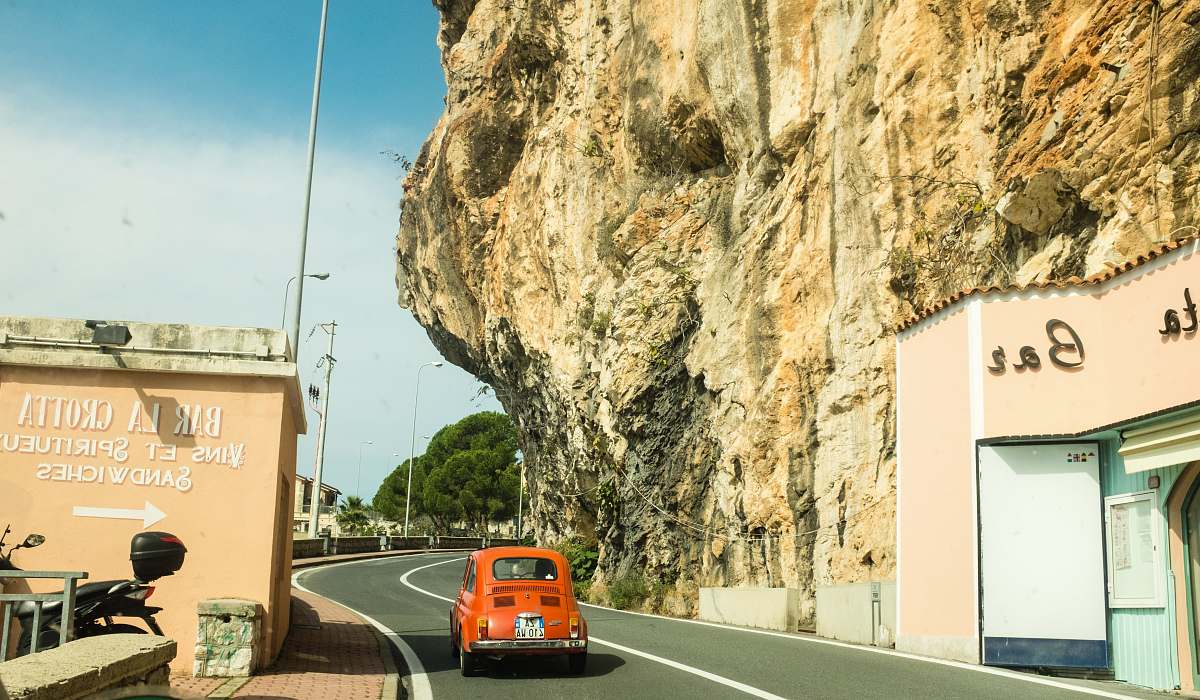 italy red Volkswagen Beetle coupe on black concrete road between concrete buildings under white and blue sky at daytime road
