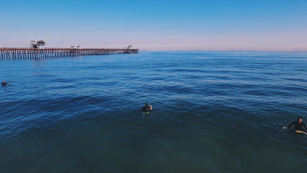 Witness the serene beauty of a vibrant blue sky illuminating three swimmers enjoying a refreshing dip in the ocean. The scene features a man swimming gracefully with a picturesque pier in the background, capturing the essence of summer relaxation and adventure. This high-quality image is perfect for travel blogs, beach resort promotions, or any content focused on aquatic activities and leisure.