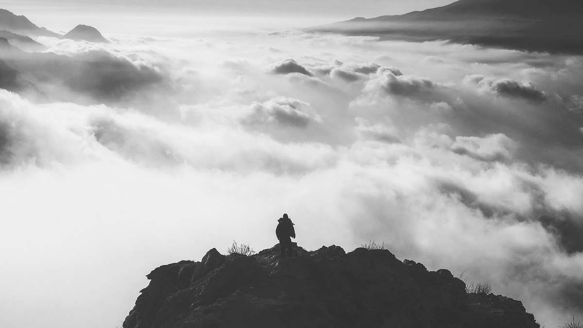 grey person standing at the edge of a mountain facing clouds during day mountain