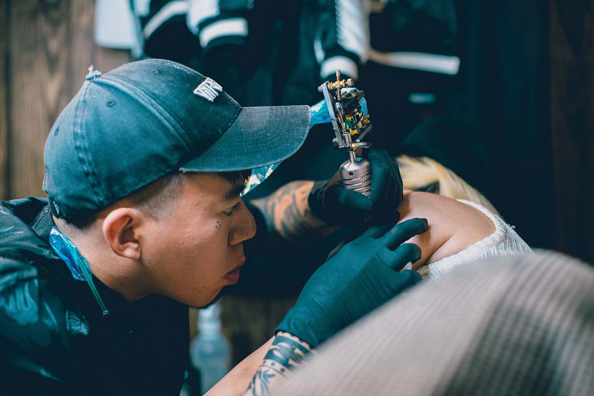 clothing man holding gray rotary tattoo machine making tattoo on person arm hat