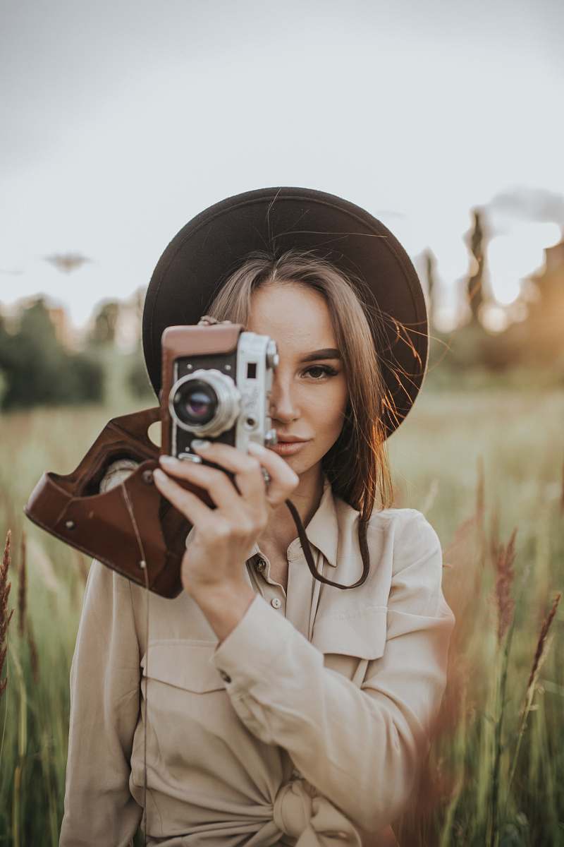 A joyful woman holds a camera, capturing her own reflection in a picturesque field bathed in daylight. This vibrant scene showcases the essence of self-expression and the beauty of nature, making it perfect for personal blogs, travel websites, or social media campaigns focused on photography, outdoor adventures, and women empowerment.