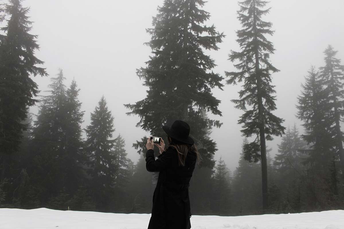 black-and-white woman takes photo at the snowfield surrounded with trees tree