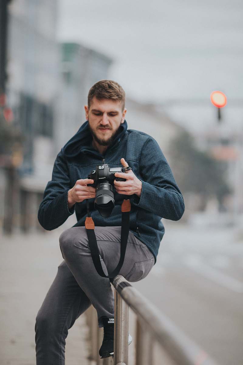 person man sitting on gray railing while holding DSLR camera during daytime photographer