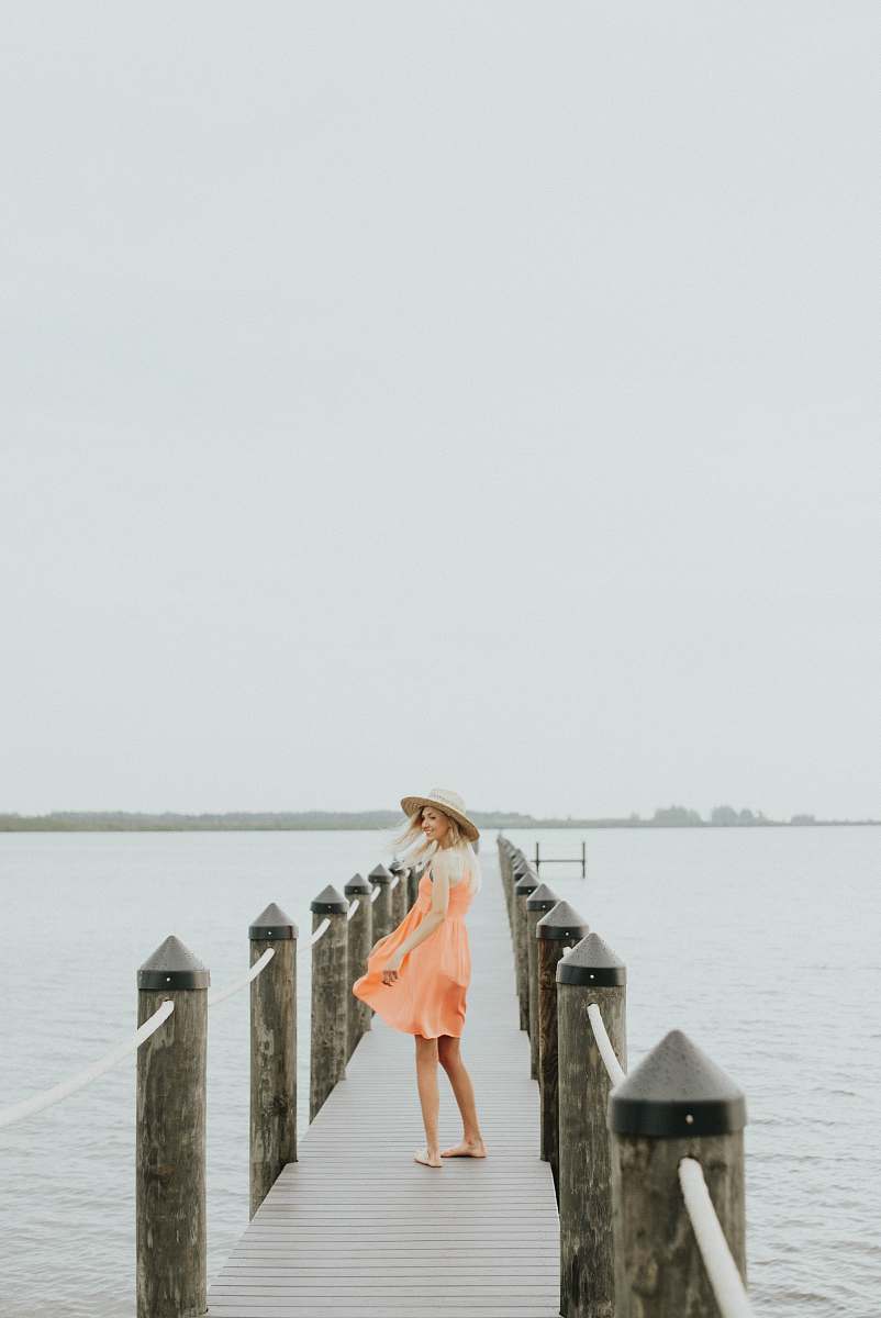 people woman in orange spaghetti strap mini dress standing on brown wooden dock during daytime woman