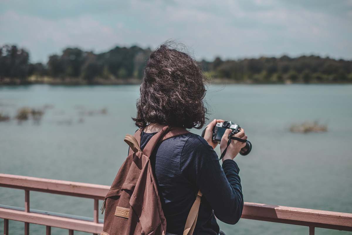 person woman wearing black shirt and brown backpack holding camera standing near railings near body of water photographer