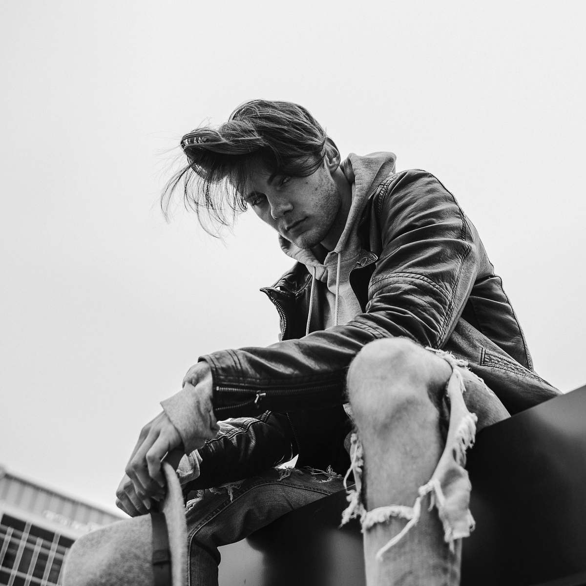 person grayscale photo of man swearing leather jacket sitting on floor near building black-and-white