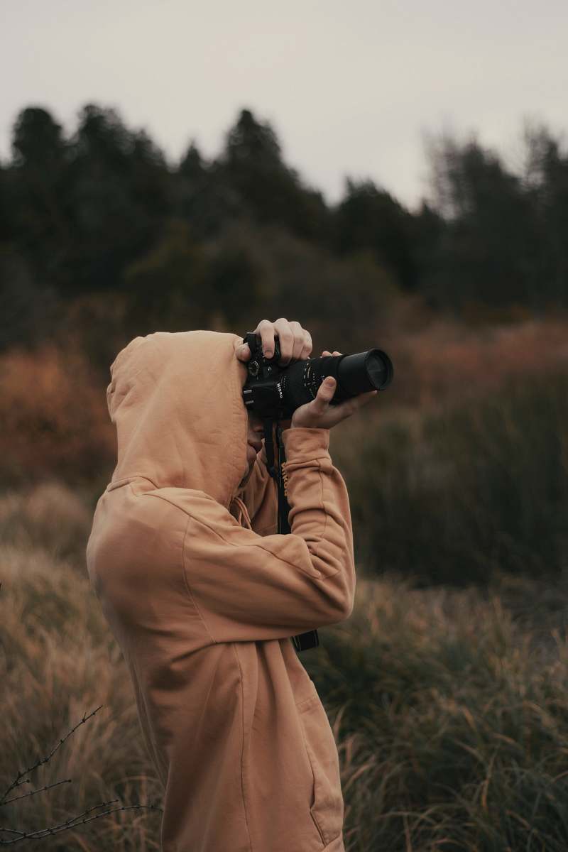 A captivating image of a woman in a hoodie, engrossed in photography with her DSLR camera, set against the serene backdrop of a lush field. This scene embodies the spirit of adventure and creativity, perfect for illustrating themes of exploration, nature, and personal expression. Ideal for use in articles about photography, outdoor activities, or lifestyle blogs.