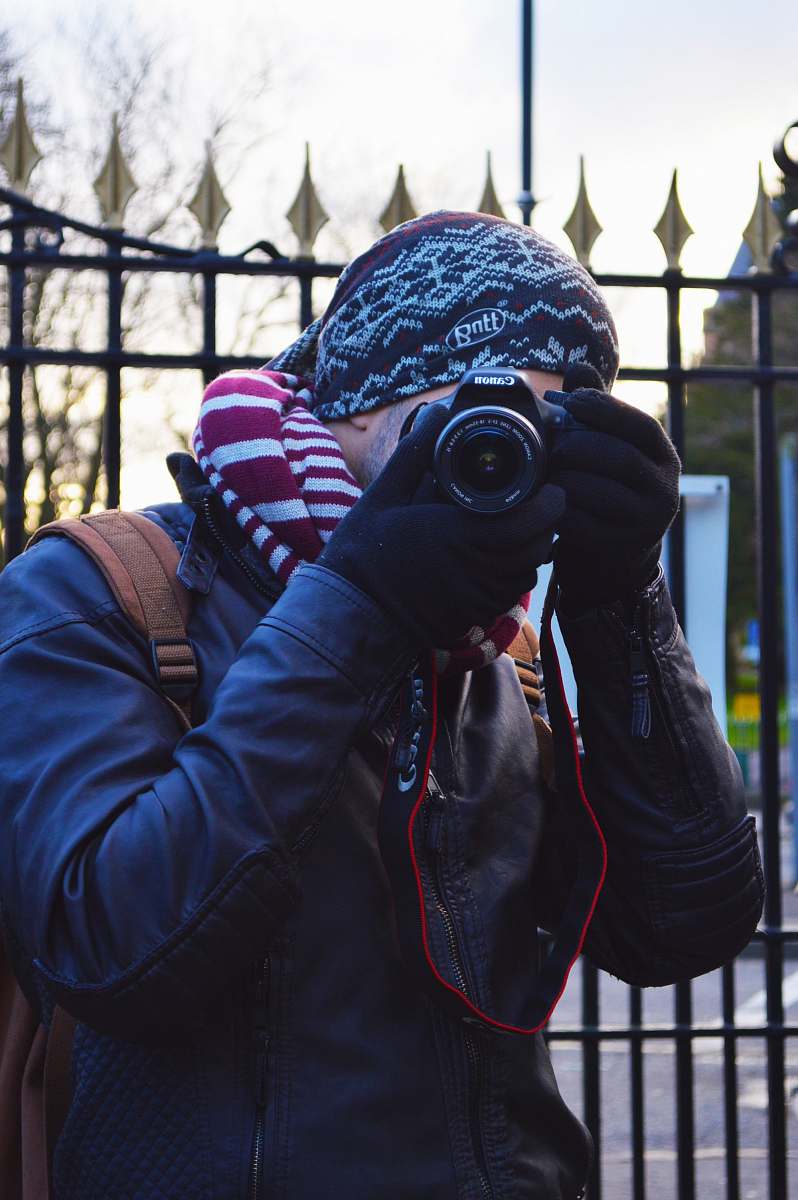 photographer man wearing black jacket using black Canon DSLR camera near black metal gate apparel