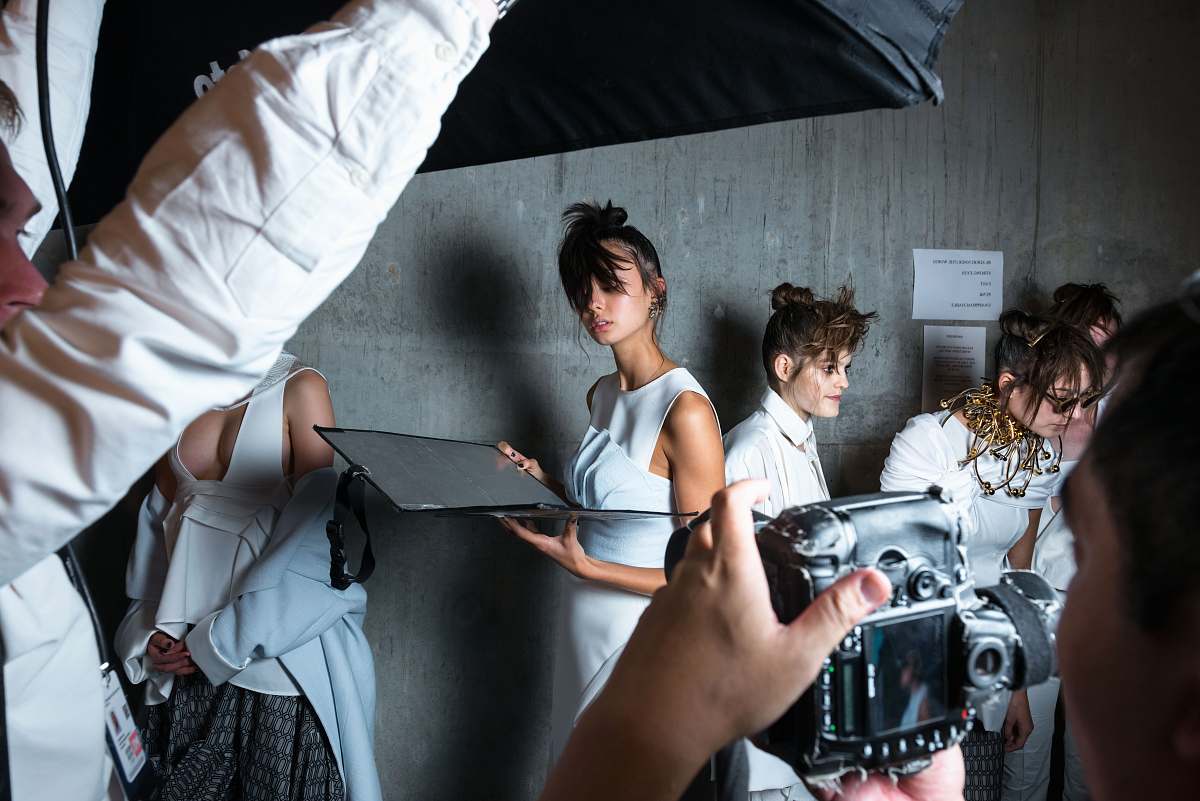 human person holding camera taking photo of woman standing near gray wall fashion
