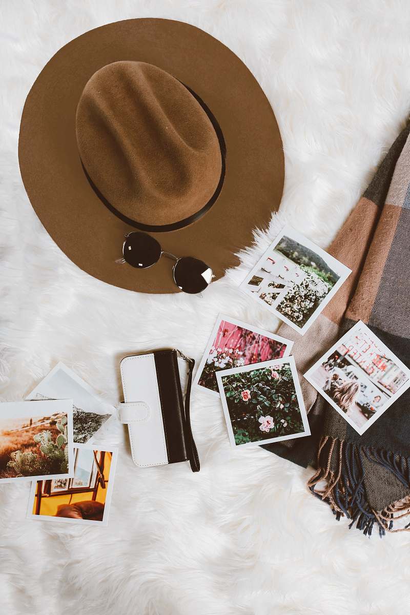 art gold-colored framed sunglasses on brown hat beside white and black leather wristlet surrounded by photos on white and gray floral textile hipster