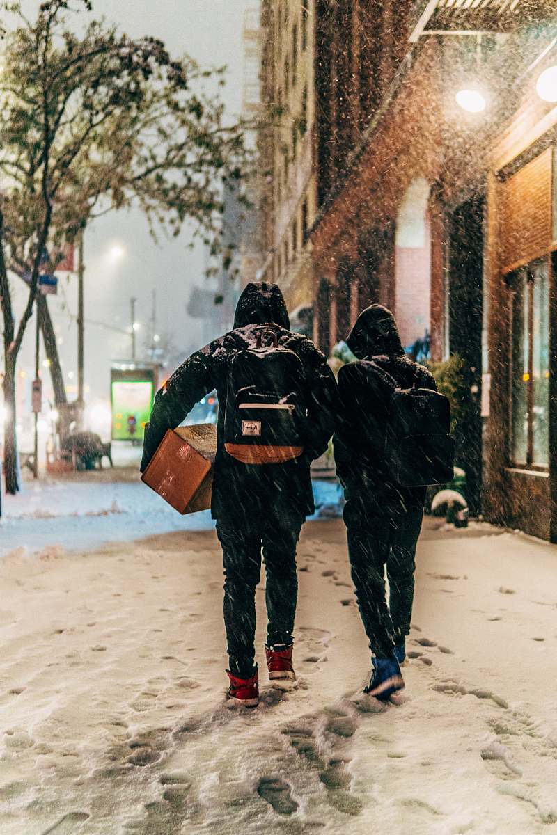 winter two people walking beside brown concrete building during snow storm