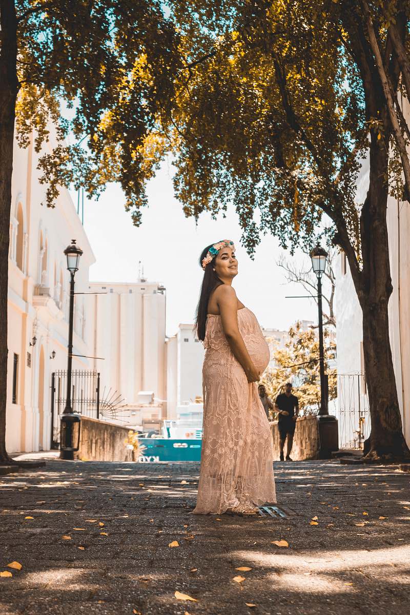 clothing smiling pregnant woman wearing beige tube dress touching her tummy while standing on concrete pathway evening dress