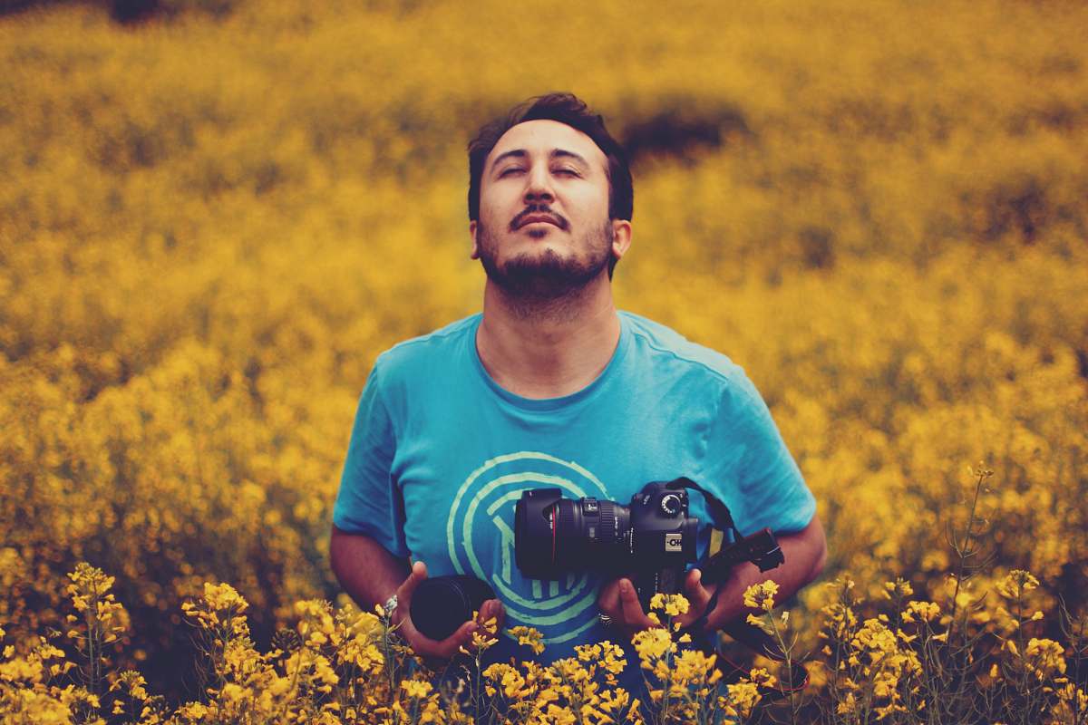 person man wearing gray crew-neck shirt holding black DSLR camera standing in the middle of yellow flowers photo