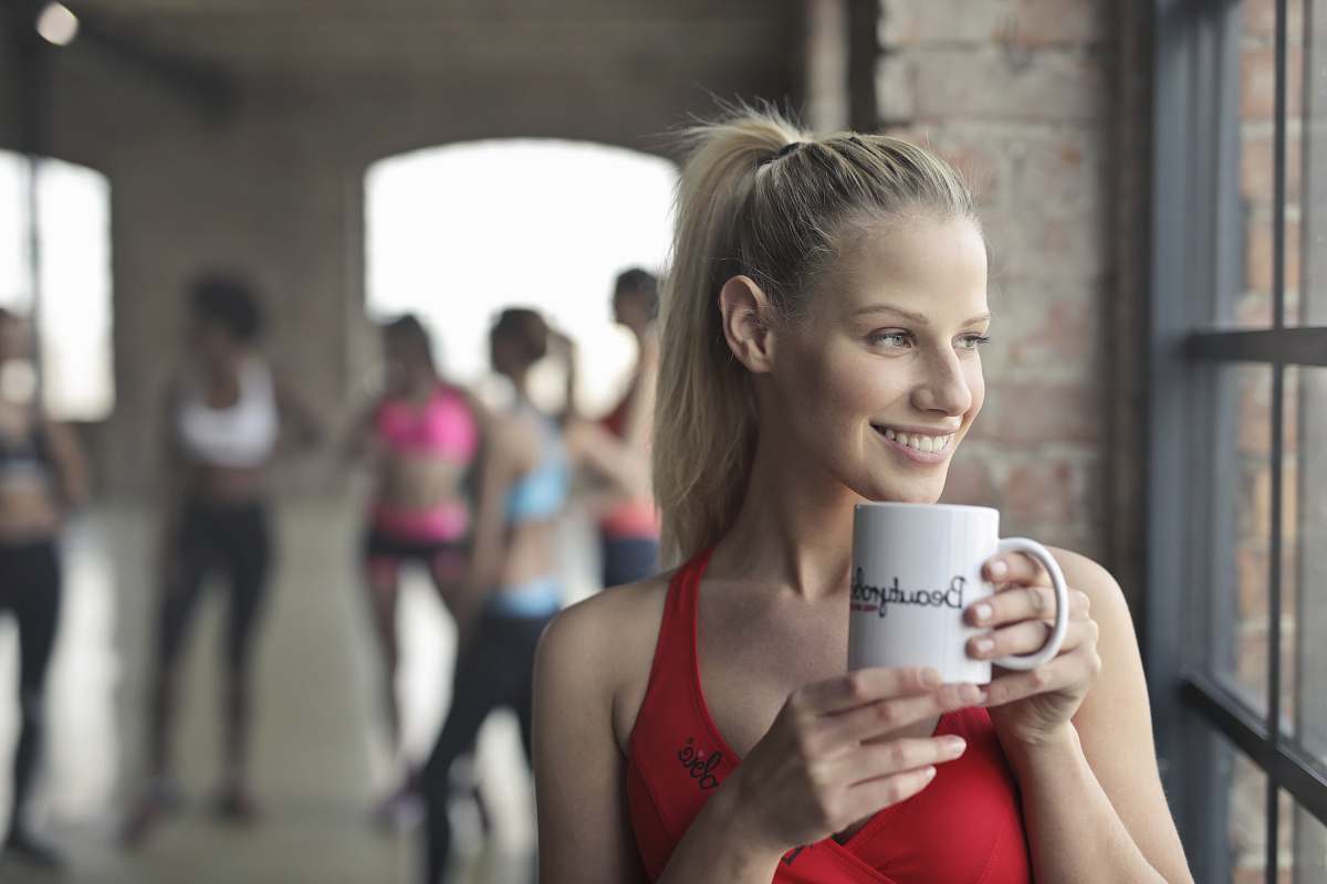 person woman holding white ceramic mug while smiling near glass window human