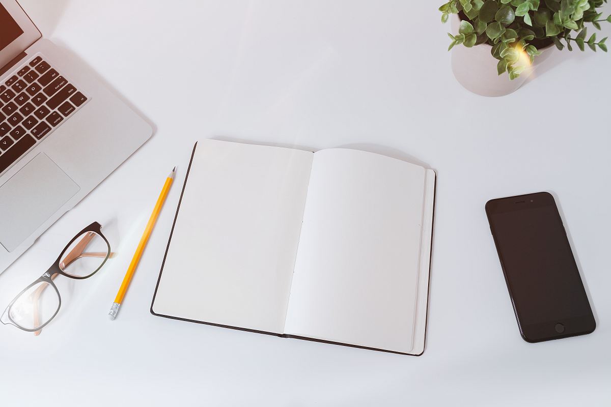 An open empty notebook on a white desk next to an iPhone and a MacBook