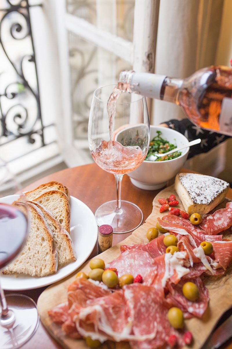 person pouring beverage on clear long-stem wine glass beside white ceramic plate with sliced bread and brown wooden board with bacons