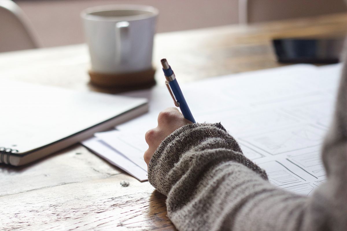 person writing on brown wooden table near white ceramic mug