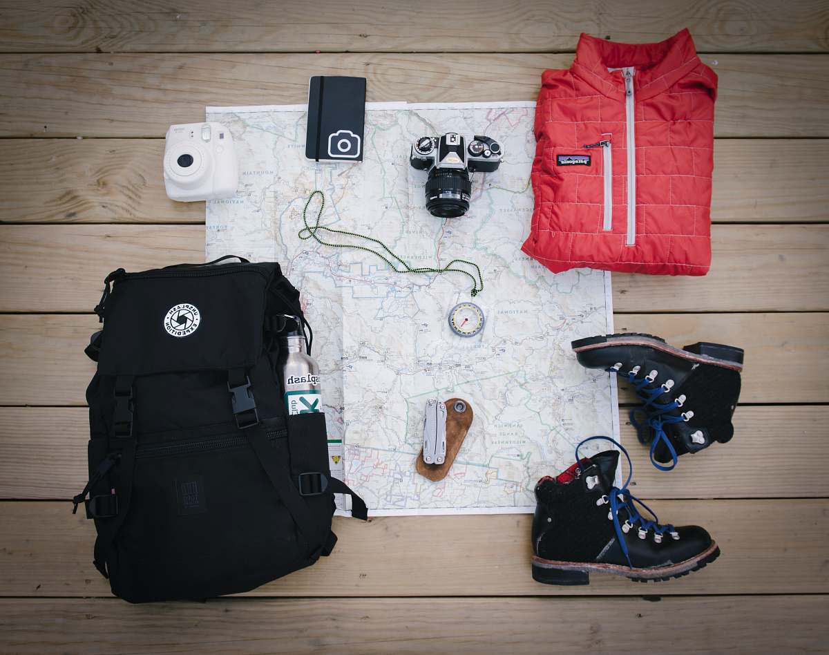 person black hiking backpack near white Fujifilm instax mini camera near black leather boots, red half-zip jacket, gray pocket watch on white map people