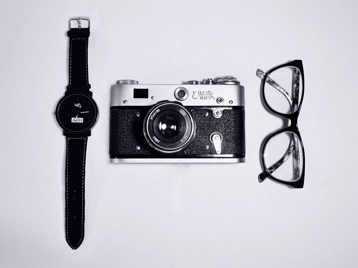 black and silver camera; eyeglasses with black frames; black analog watch