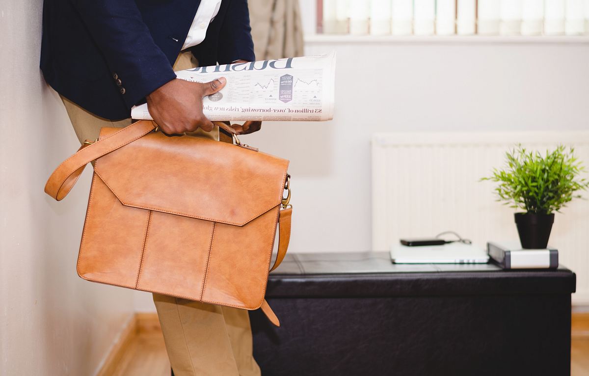 man holding brown leather bag leaning on white wall