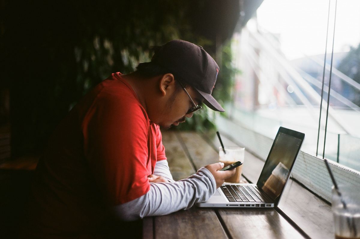 man holding phone in with laptop beside cup